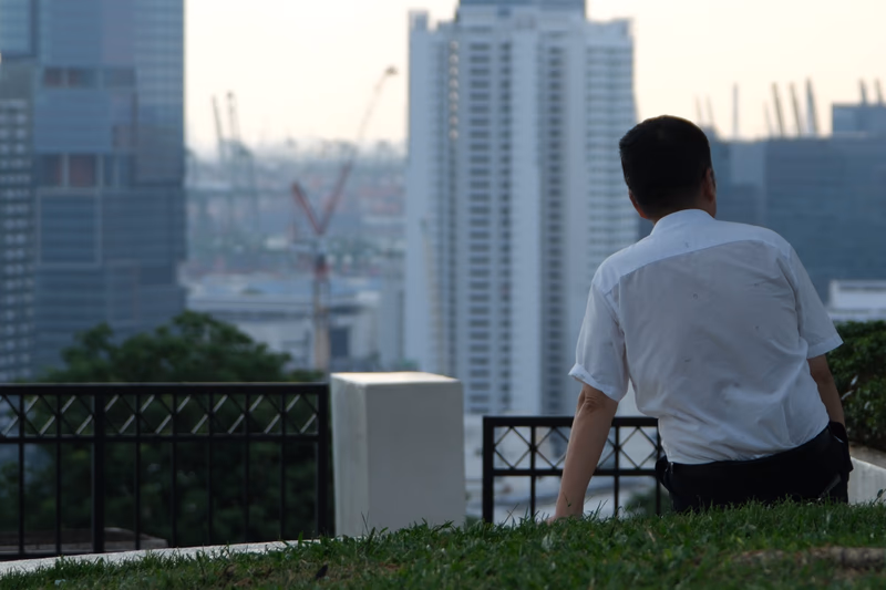 A man sitting on grass in a city with a view of skyscrapers and cranes.