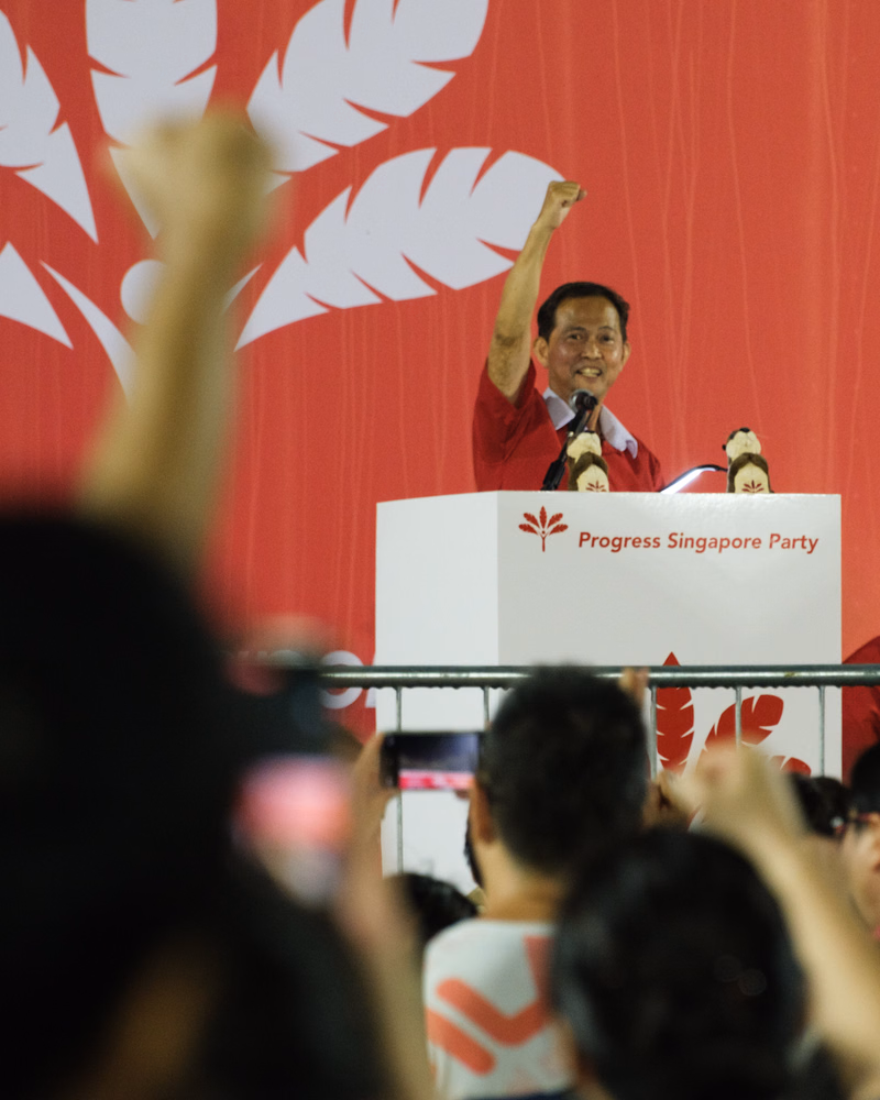 A politician is speaking at a rally on a stage with a large red background featuring white leaf shapes, and a sign reads "Progress Singapore Party".