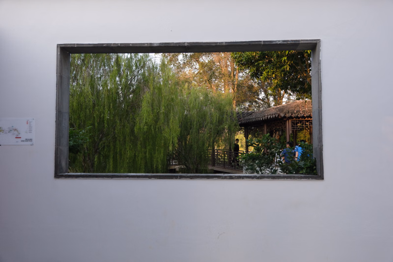 A rectangular window frames a view of lush weeping willow trees next to a traditional wooden pavilion in a garden setting.