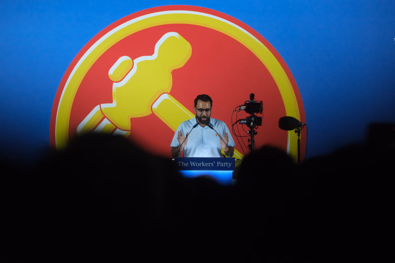 A man stands on a stage in front of a large red and yellow sign that reads "The Workers' Party".