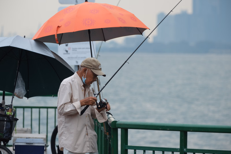 An older fisherman wearing a cap and mask fishes off a pier under an orange umbrella, with a city skyline visible in the background.