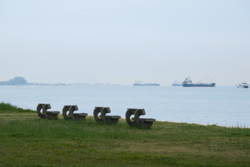 A row of concrete benches sits on a grassy bank overlooking a calm body of water with several ships in the distance.