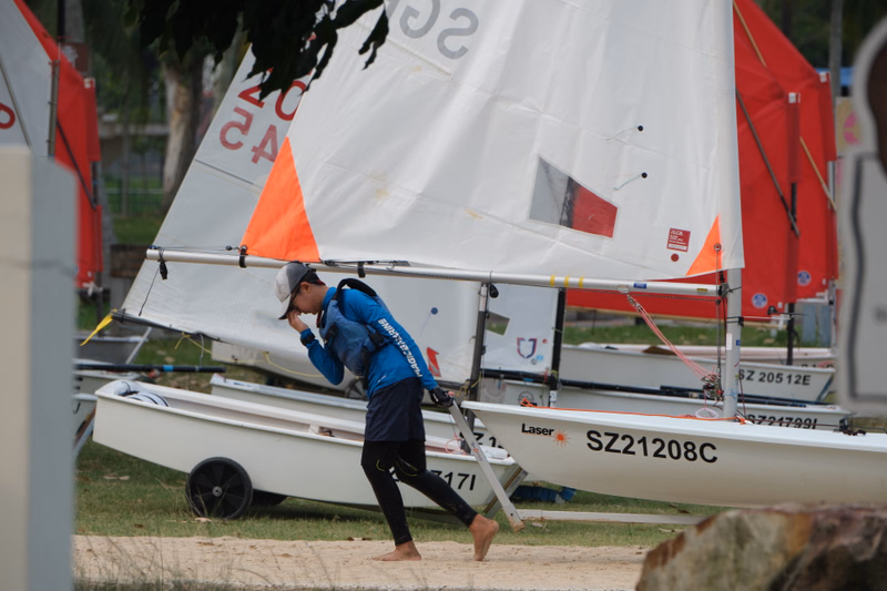 A sailor in a blue top is near a large white sailboat with orange accents, which has the text "SZ21208C" visible.
