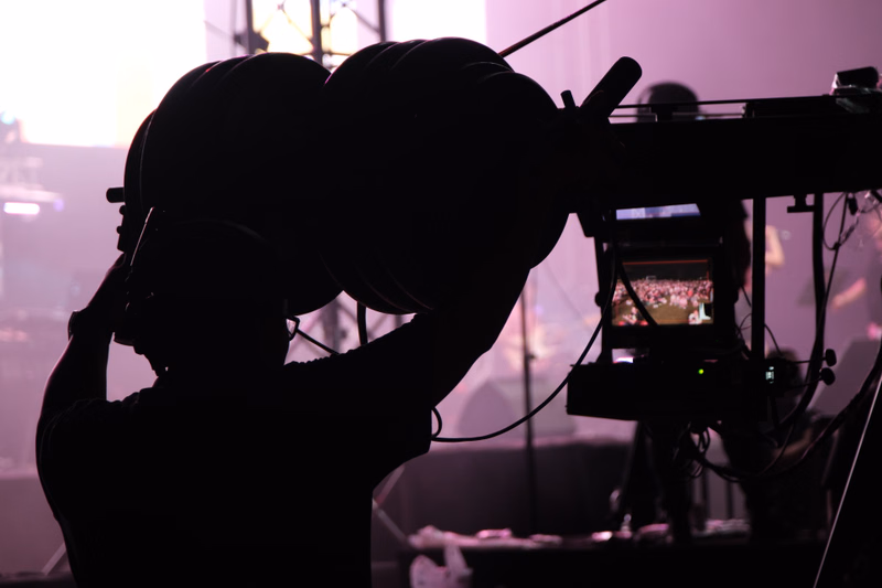 A videographer in silhouette holds a large camera setup while filming in a dimly lit studio environment.