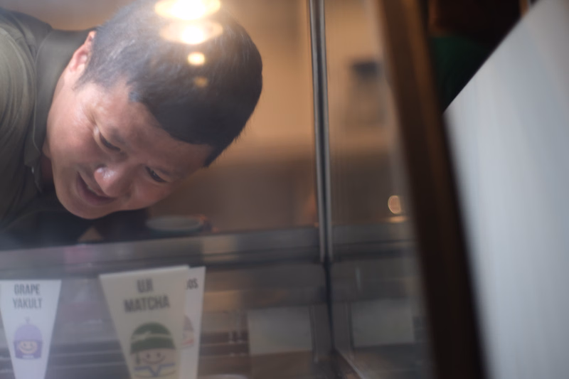 A man looks into a refrigerated display case showing signs for "GRAPE YAKULT" and "UJI MATCHA".