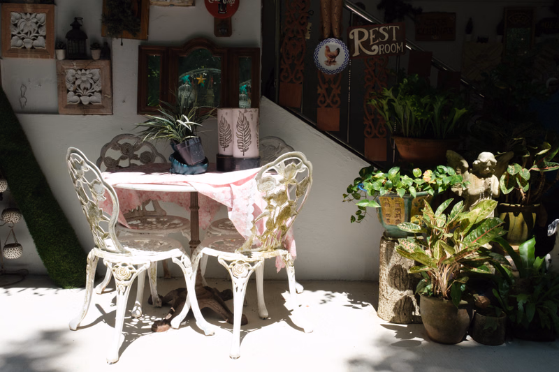 A set of white, ornate patio chairs surrounds a small table decorated with a pink cloth, set against a wall with a "REST ROOM" sign.