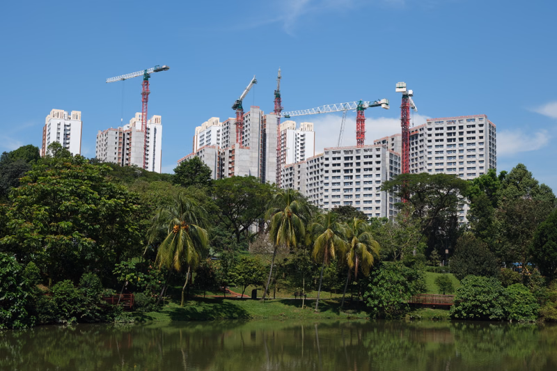 Modern apartment buildings under construction with cranes are visible behind a calm lake surrounded by lush tropical trees.