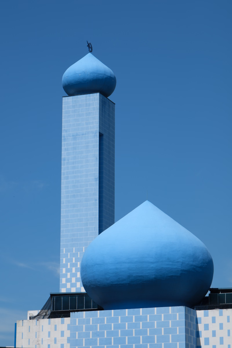 A modern skyscraper with a blue dome on top stands next to a large, smooth blue dome structure against a clear blue sky.