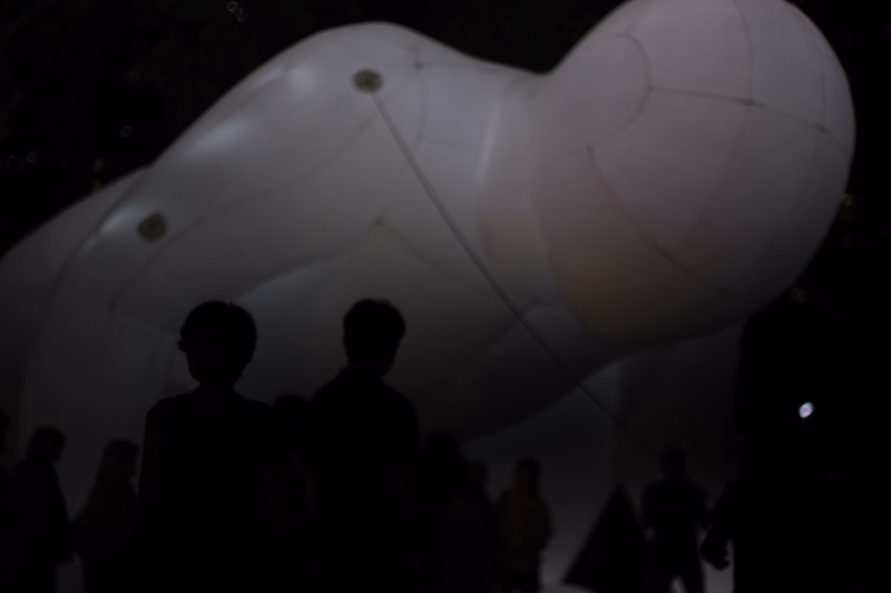 A large, white inflatable sculpture dominates the frame above a crowd of silhouetted people at night.