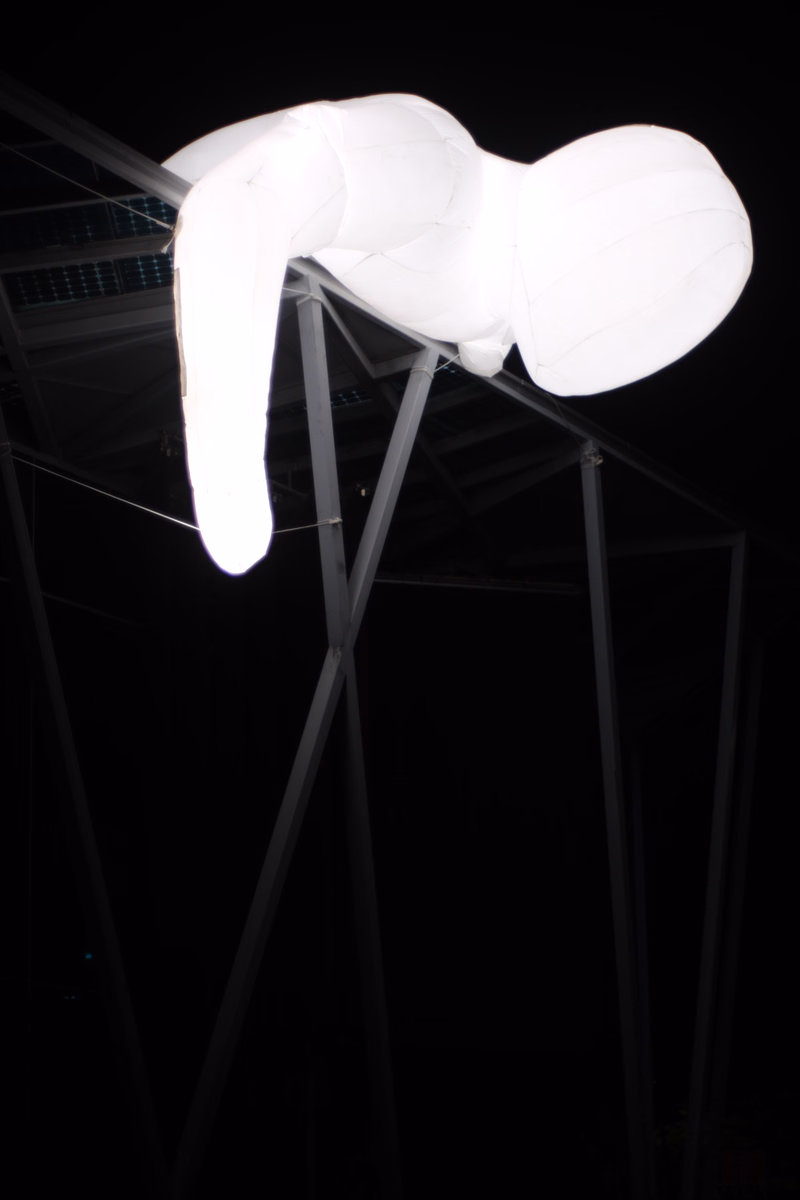A large, white, inflated balloon shaped like a stylized animal or figure hangs suspended beneath a metal framework against a black background.