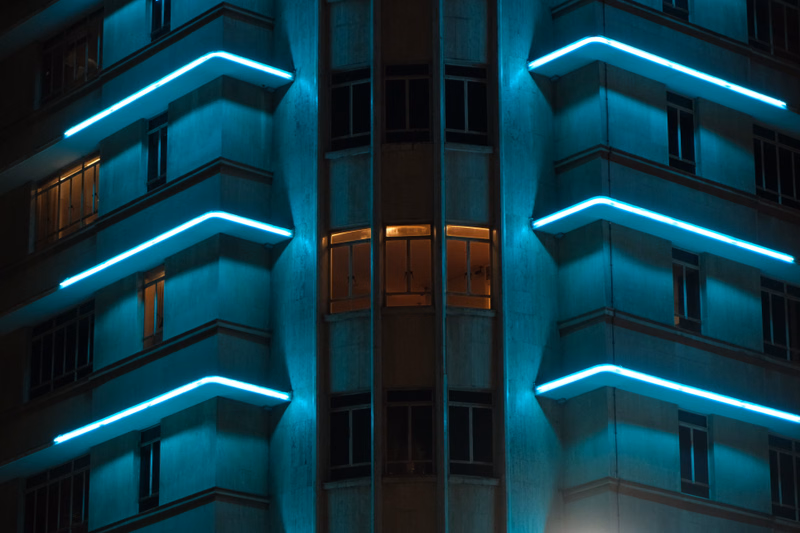 A modern building facade at night is illuminated by bright, glowing blue neon strip lights outlining its architectural features.