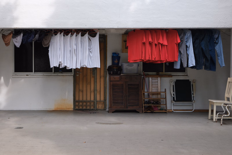 The open front of a beach hut displays hanging laundry, including white shirts and red garments, next to a wooden cabinet.