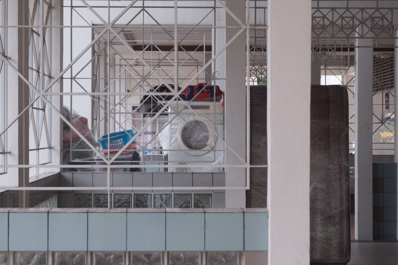 A view through a white, geometric metal grate shows several washing machines and laundry items inside a bright, industrial-looking space.