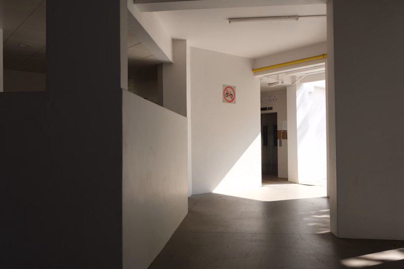 A bright interior hallway with white walls and a doorway leading to another brightly lit area, featuring a red and white 'No' sign on the wall.