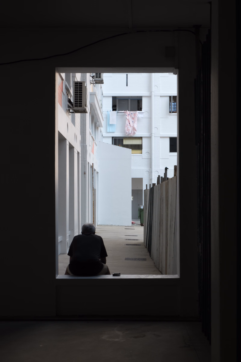 A person sits on the ground in a narrow, bright alleyway lined with white buildings, with clothes hanging to dry.