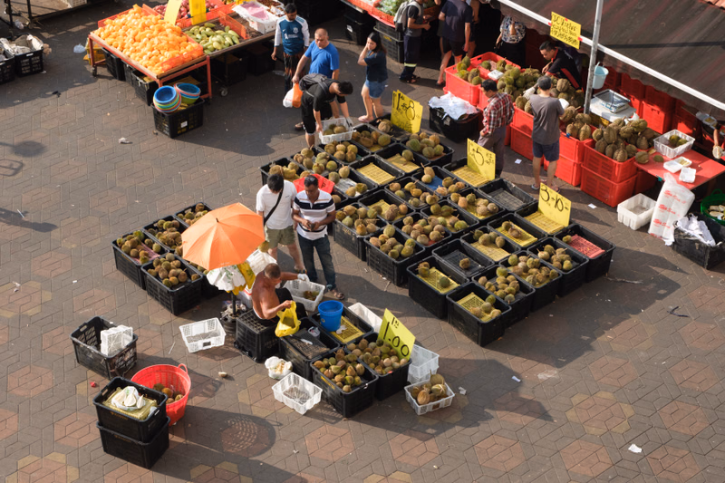 A busy outdoor fruit stall displays various types of produce, including oranges and green fruits, with several people browsing the market.