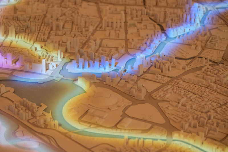 A detailed architectural model shows a city layout with a winding river flowing through the urban development.