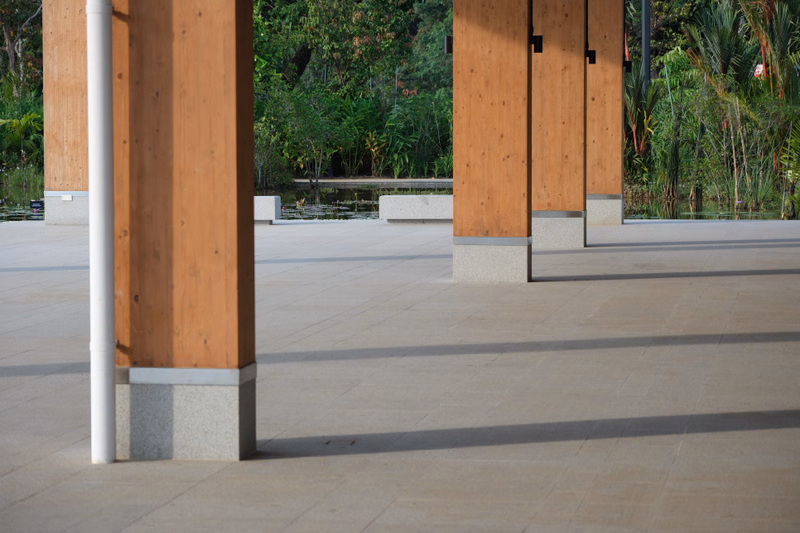 A row of tall, wooden pillars with stone bases lines a wide, paved outdoor walkway surrounded by lush tropical foliage.