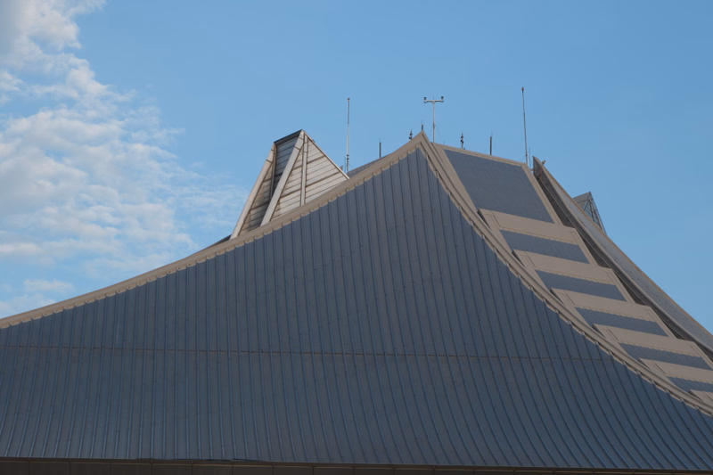A large, steeply pitched metal roof of a modern building is visible against a bright blue sky with some clouds.