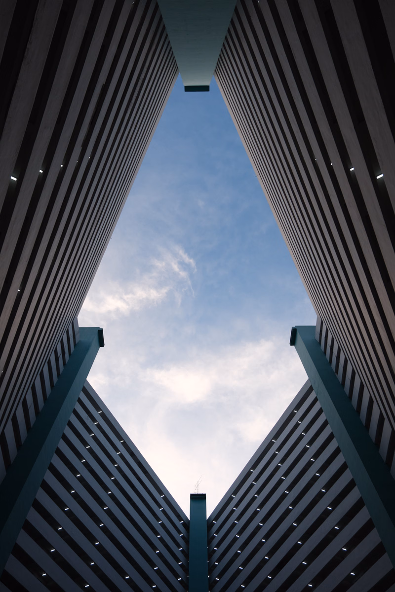 A low-angle view looking up through the vertical slats of a modern building facade against a cloudy blue sky.