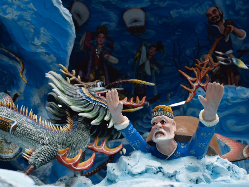 A detailed sculpture features a fierce dragon next to a man with raised arms, set against a backdrop of other fantasy figures.