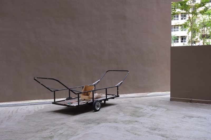 A black metal utility cart with a wooden base sits on a bare concrete floor against a large brown wall.