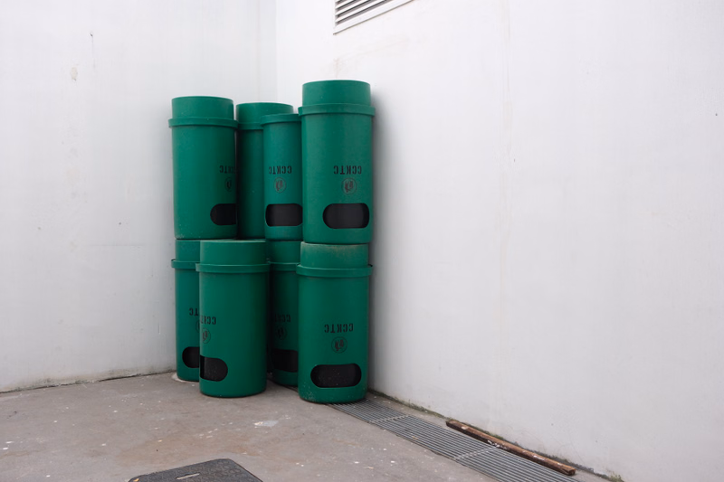 A row of several green trash and recycling bins are lined up against a white wall outdoors.