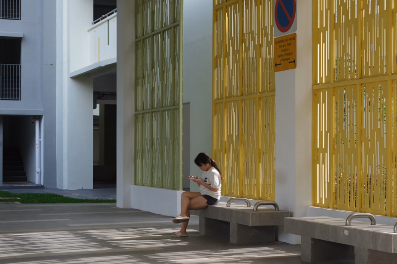 A young woman sits on a concrete bench next to a yellow slatted screen outside a modern white building.