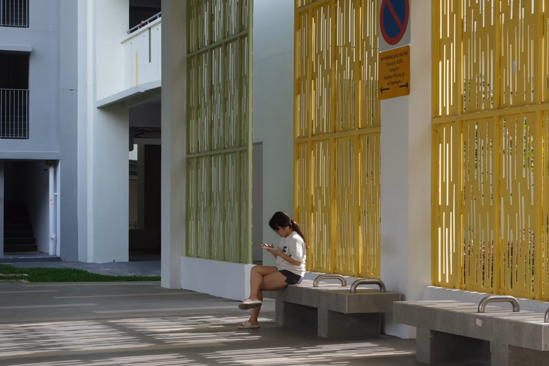 A woman sitting on a bench in front of a modern building with a yellow wall and a no parking sign.