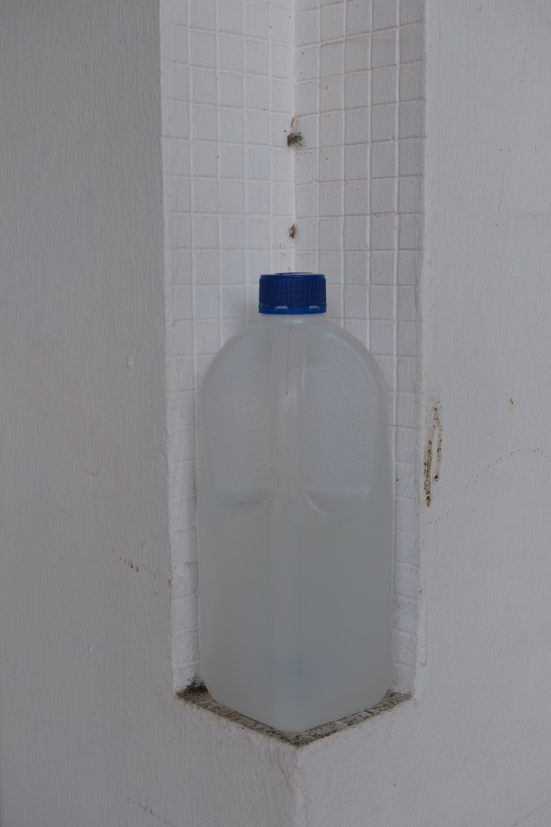 A translucent plastic water bottle with a blue cap is stored in a recessed niche in a white tiled wall.