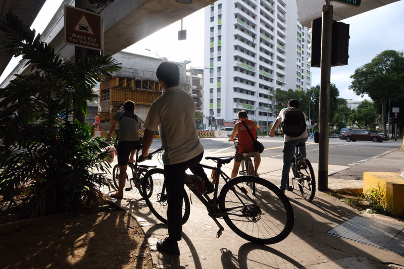 A man in a light shirt rides a bicycle on a sidewalk next to a large modern high-rise building.