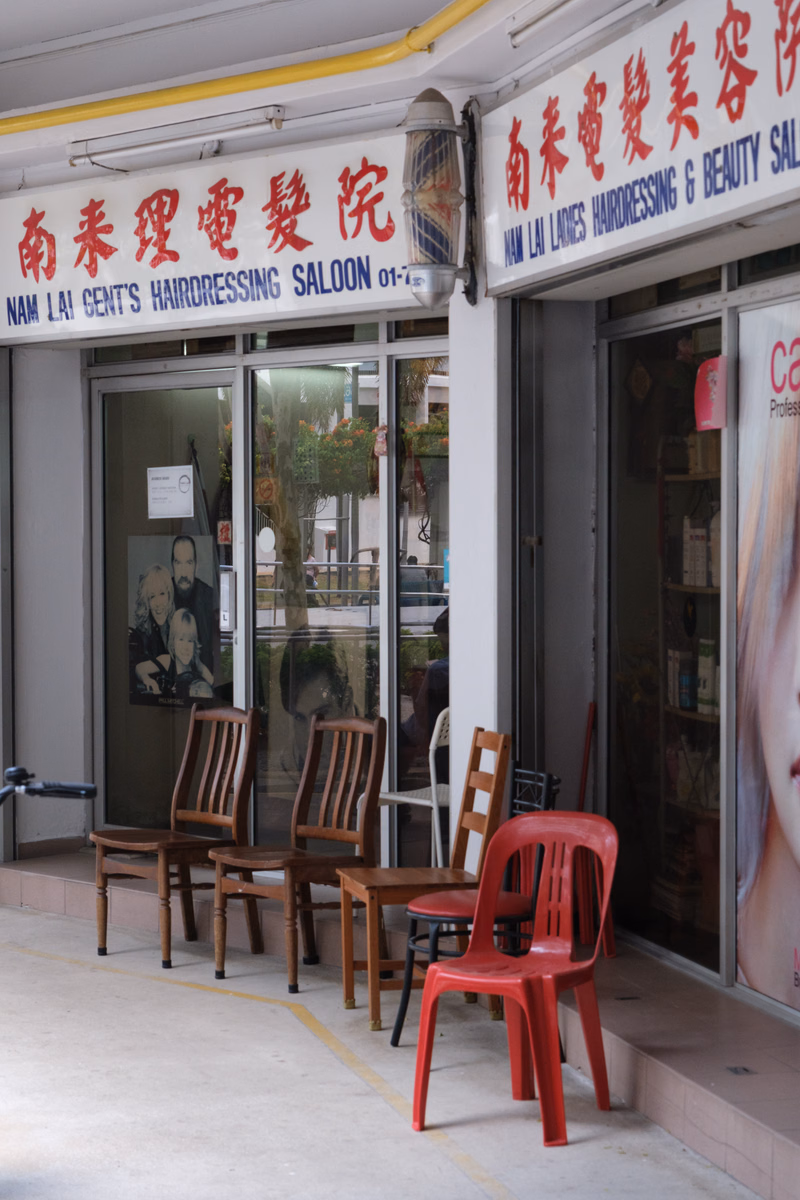 The storefront for "南來理髮院 NAM LAI GENT'S HAIRDRESSING SALOON 01-7" features several wooden and red chairs outside.