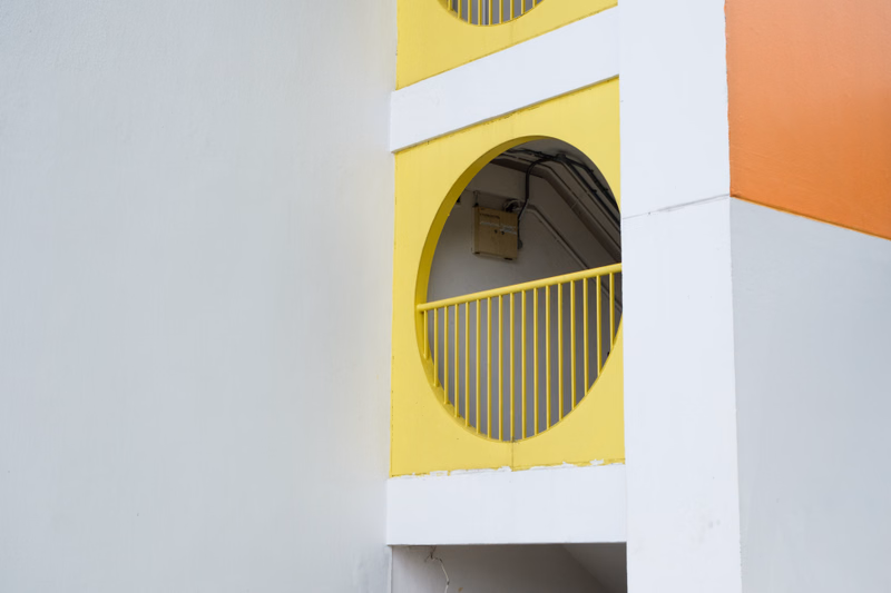 A section of a building features a circular window with a yellow railing, set against white and orange painted walls.