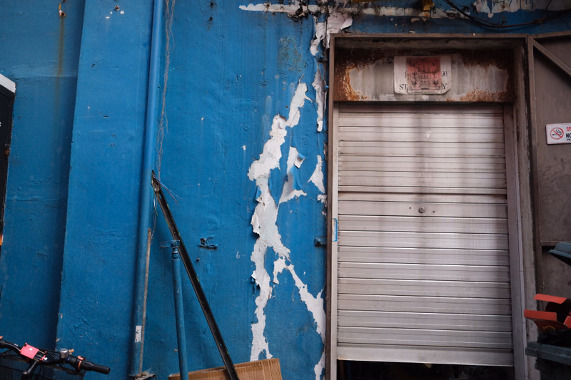 A weathered blue metal wall next to a closed silver roller shutter door, with a sign above the shutter that reads "STILL".