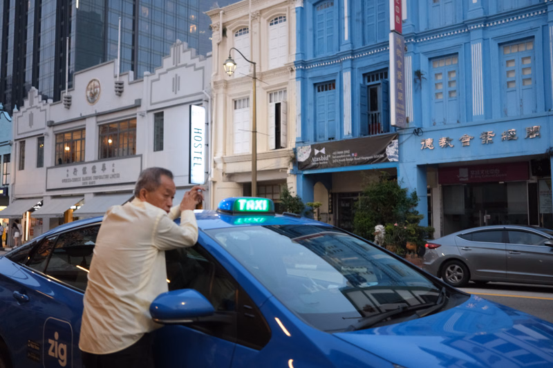 A man in a light-colored shirt stands next to a blue taxi on a city street lined with buildings, one of which has a sign reading "HOSTEL".