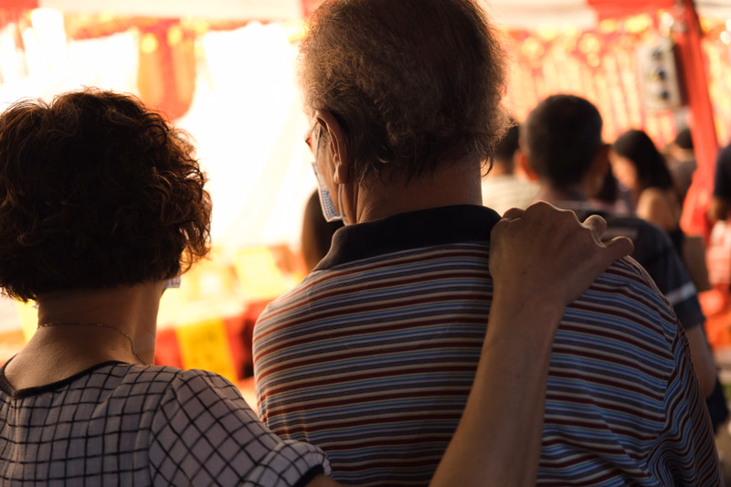 Several people are gathered outdoors in bright sunlight, with a blurred background of red structures.