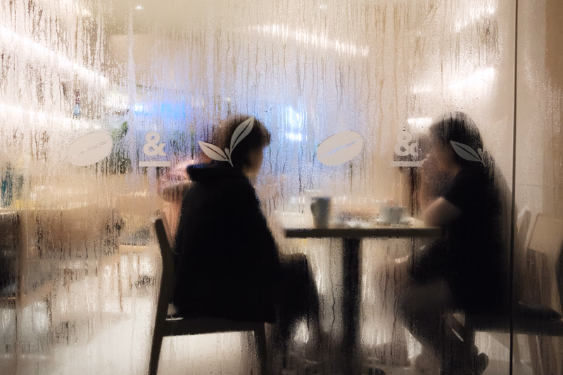 Two people sit at a table inside a cafe, viewed through a frosted window with faint '&' logos visible.