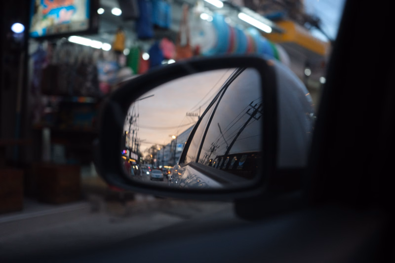 A car side mirror reflects a view of a city street with buildings and vehicles under a bright sky.