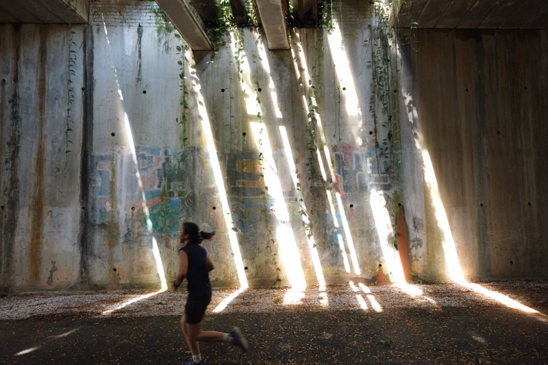 A runner exercises in an outdoor area with tall concrete walls illuminated by strong beams of sunlight.