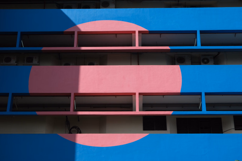 A modern building facade features a large pink circle overlaid on a bright blue background, with multiple horizontal windows visible.