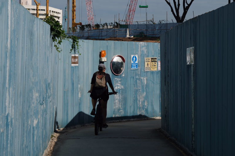 A cyclist rides down a path between tall blue fences, with construction cranes visible in the background and a sign reading "SLOW CAUTION".