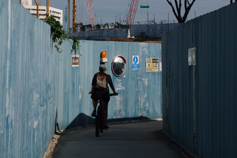 A man rides a bicycle on a narrow path between two blue fences, with construction sites and signage visible in the background.