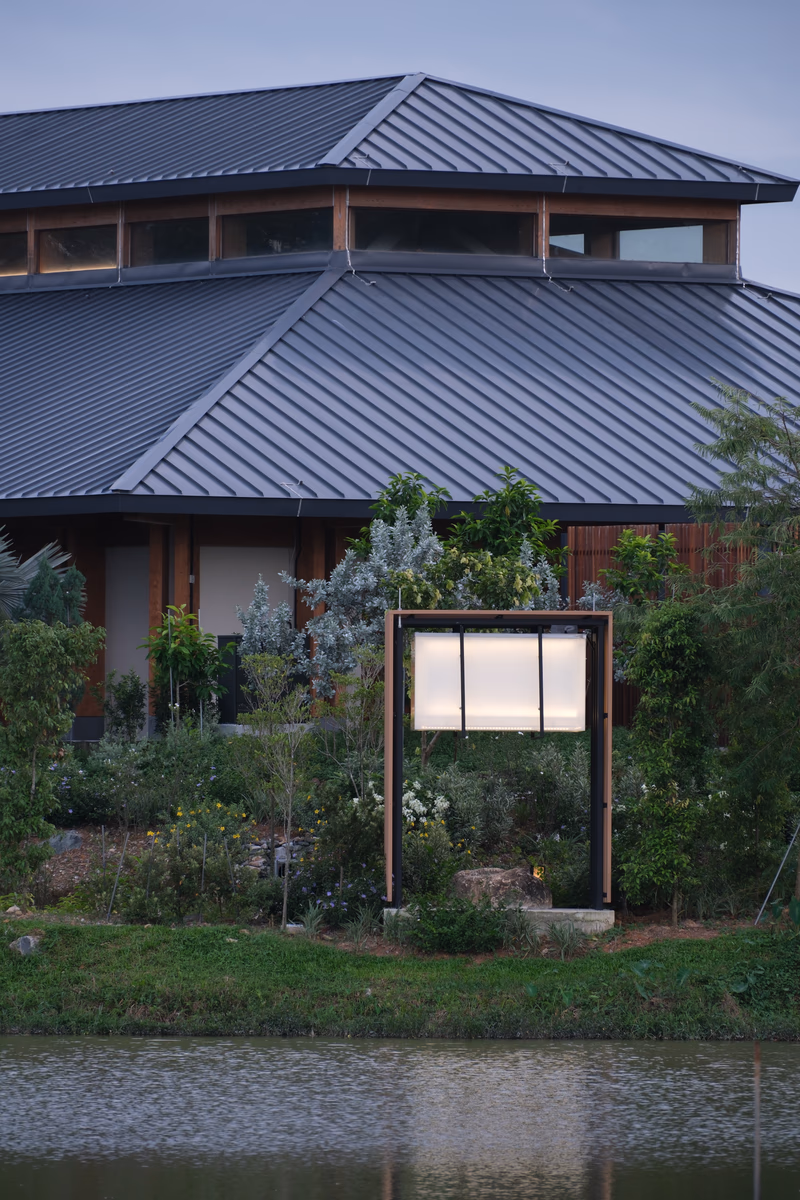 A modern building with a metal roof and large windows surrounded by lush greenery and a body of water