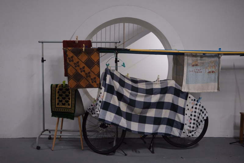 A large blue and white checkerboard quilt is displayed on a bicycle wheel inside a room with a large archway.