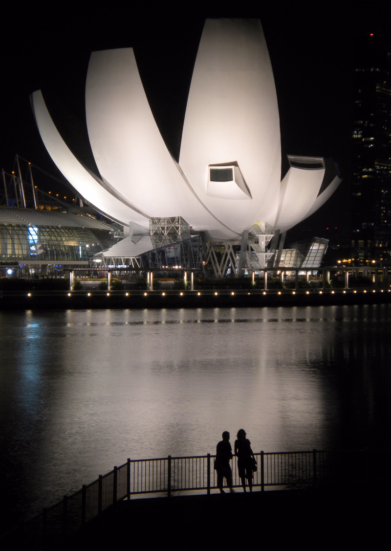 The ArtScience Museum at Marina Bay Sands is illuminated at night, with two people silhouetted on a pier overlooking the water.