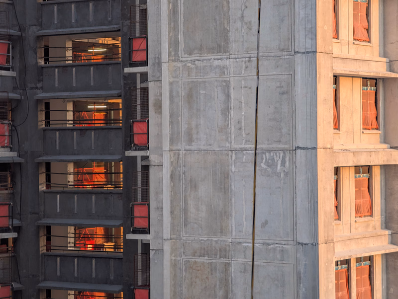 A photograph showing the textured concrete facade of a modern building next to a dark, multi-story apartment building with visible windows.