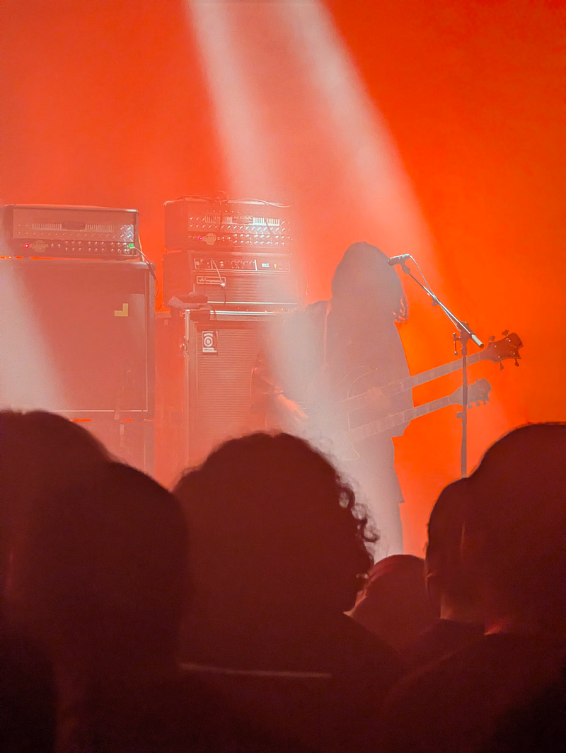 A musician plays an electric guitar on stage illuminated by bright orange spotlights in front of large amplifiers.
