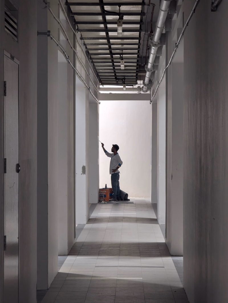 A construction worker stands in a long, empty hallway under an exposed metal ceiling, appearing to paint or work on the wall.