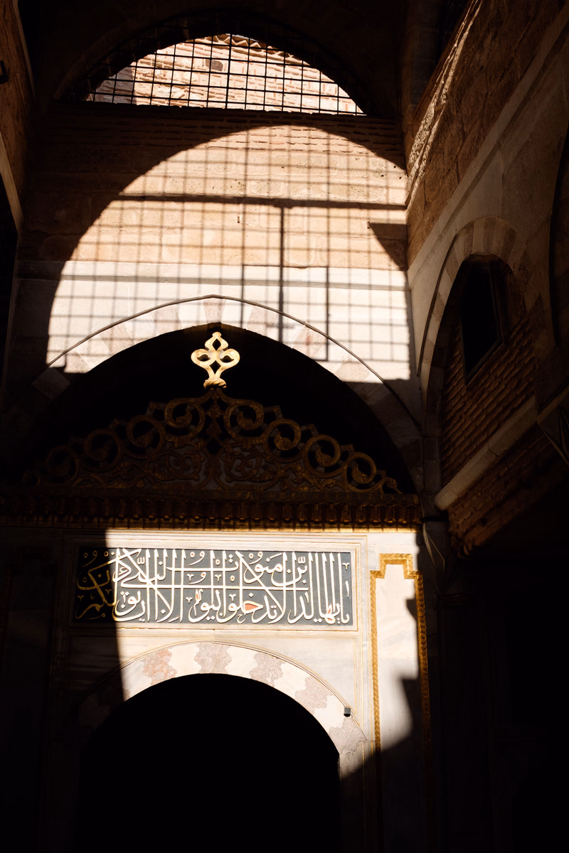 Sunlight streams through a large arched window into a mosque interior, illuminating Arabic calligraphy on a decorated wall.