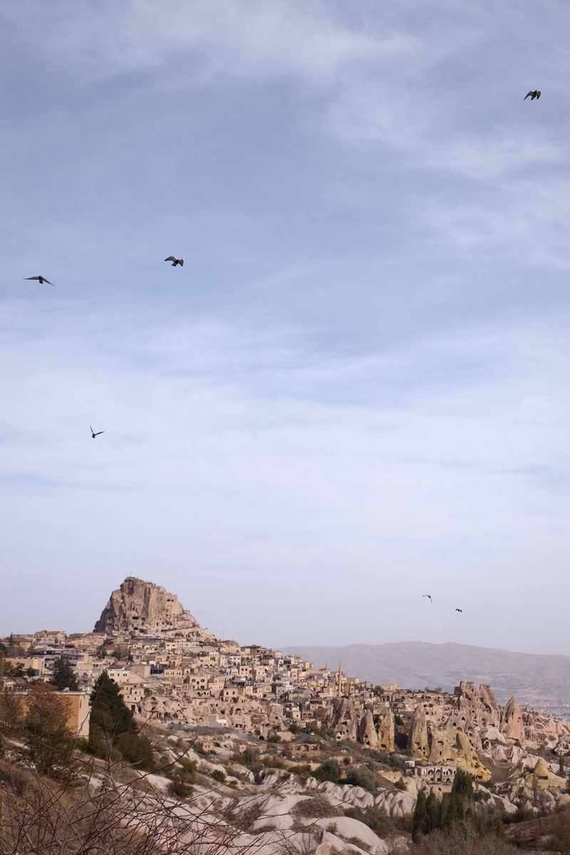 A historic town built into a rocky hillside under a vast, cloudy sky, with several birds flying overhead.